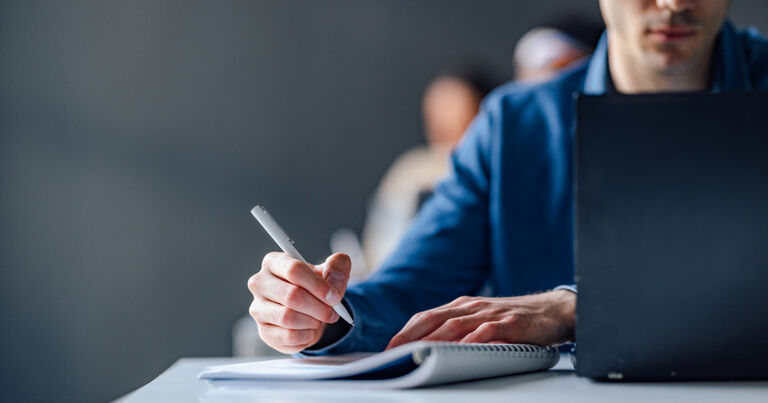student sitting at a desk with laptop and writing in a notebook